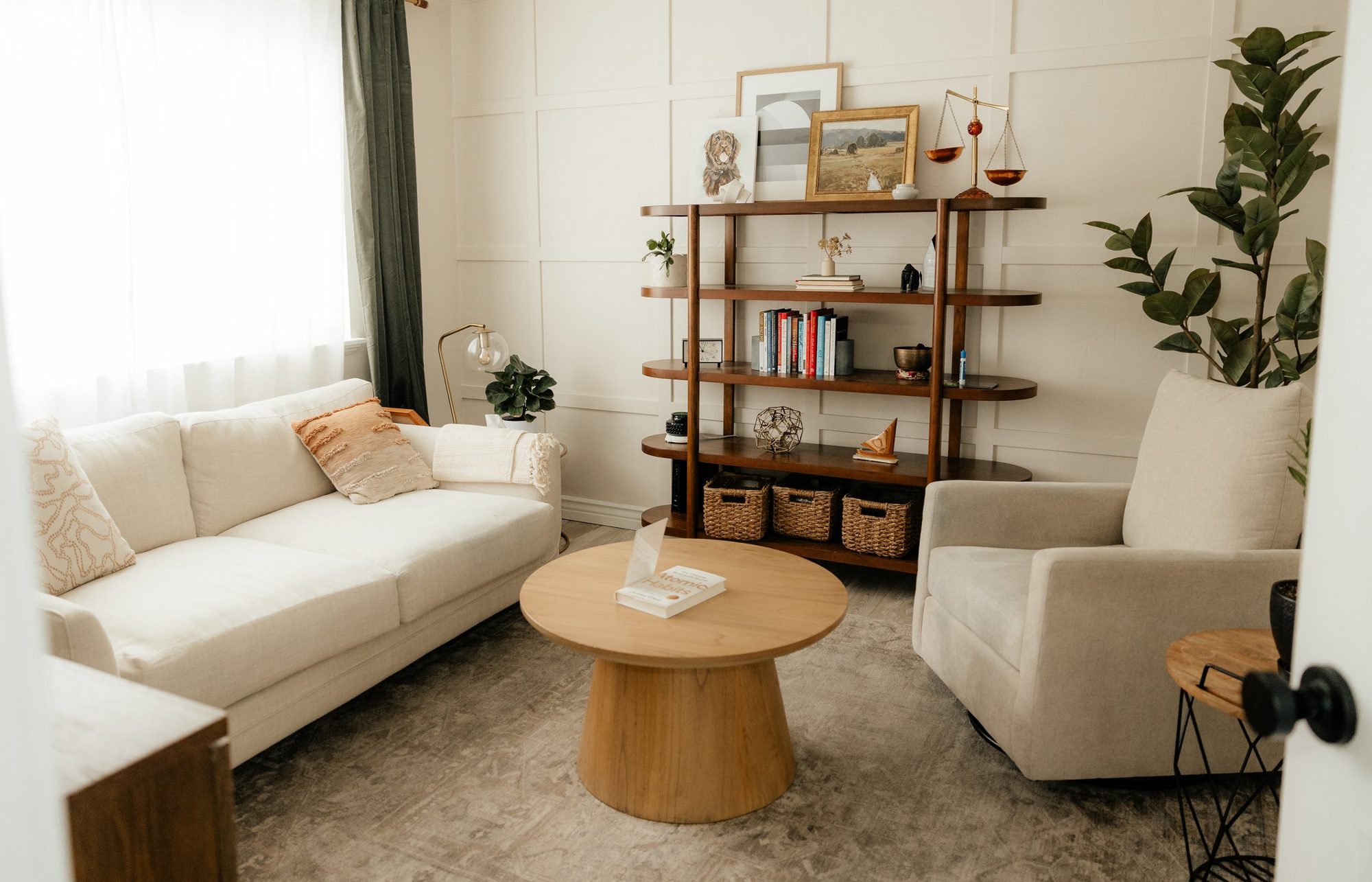A cozy, minimalist Therapy office at Tangible Therapy in Provo Utah features a white couch with cushions, a round wooden coffee table, and a shelf with books, plants, and decorative items.