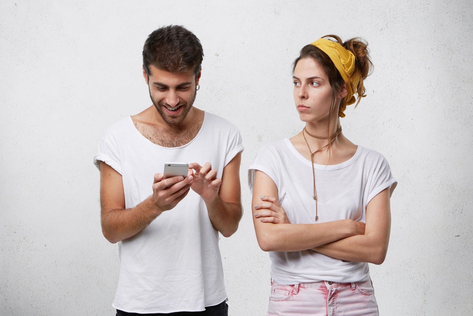 A man in a white t-shirt looks at his smartphone with a happy expression while a woman in a yellow headband stands beside him with her arms crossed, looking skeptical.