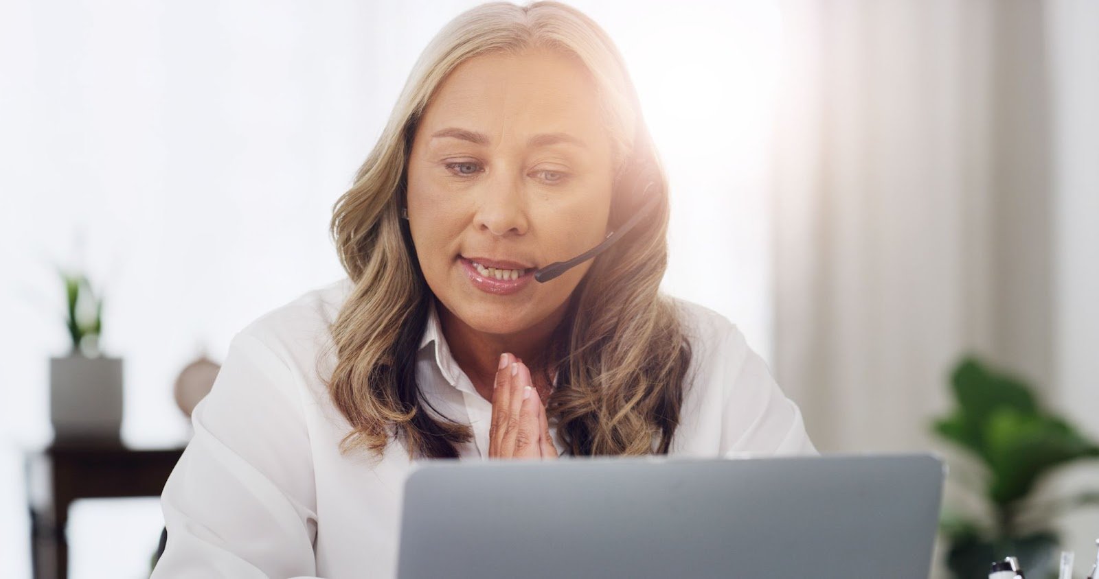 A professional woman with blonde hair wearing a headset and white blouse, speaking into her laptop during a virtual consultation or customer support call.