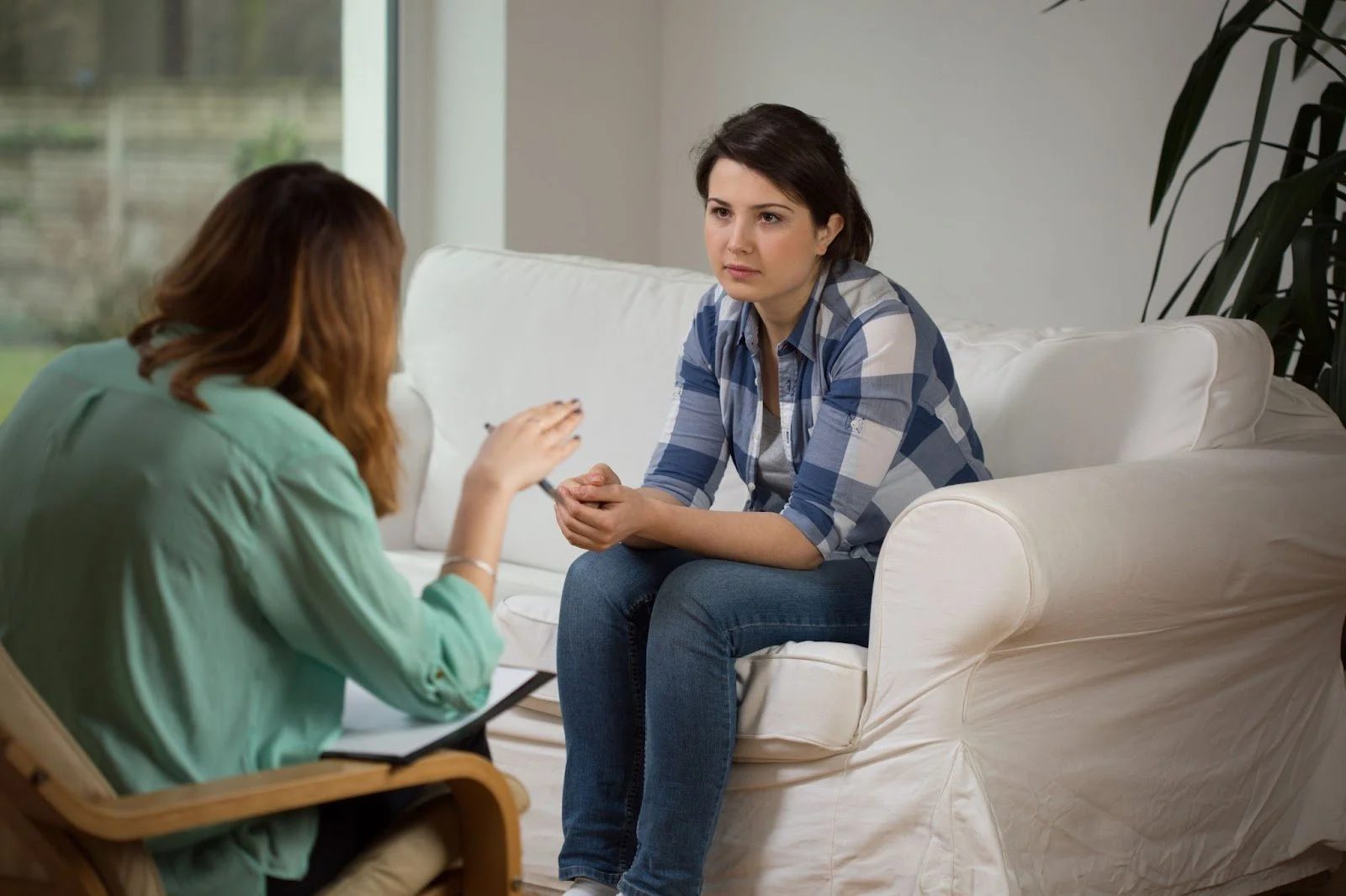 A woman in a blue-and-white plaid shirt sitting on a white sofa and listening intently to a therapist or consultant.