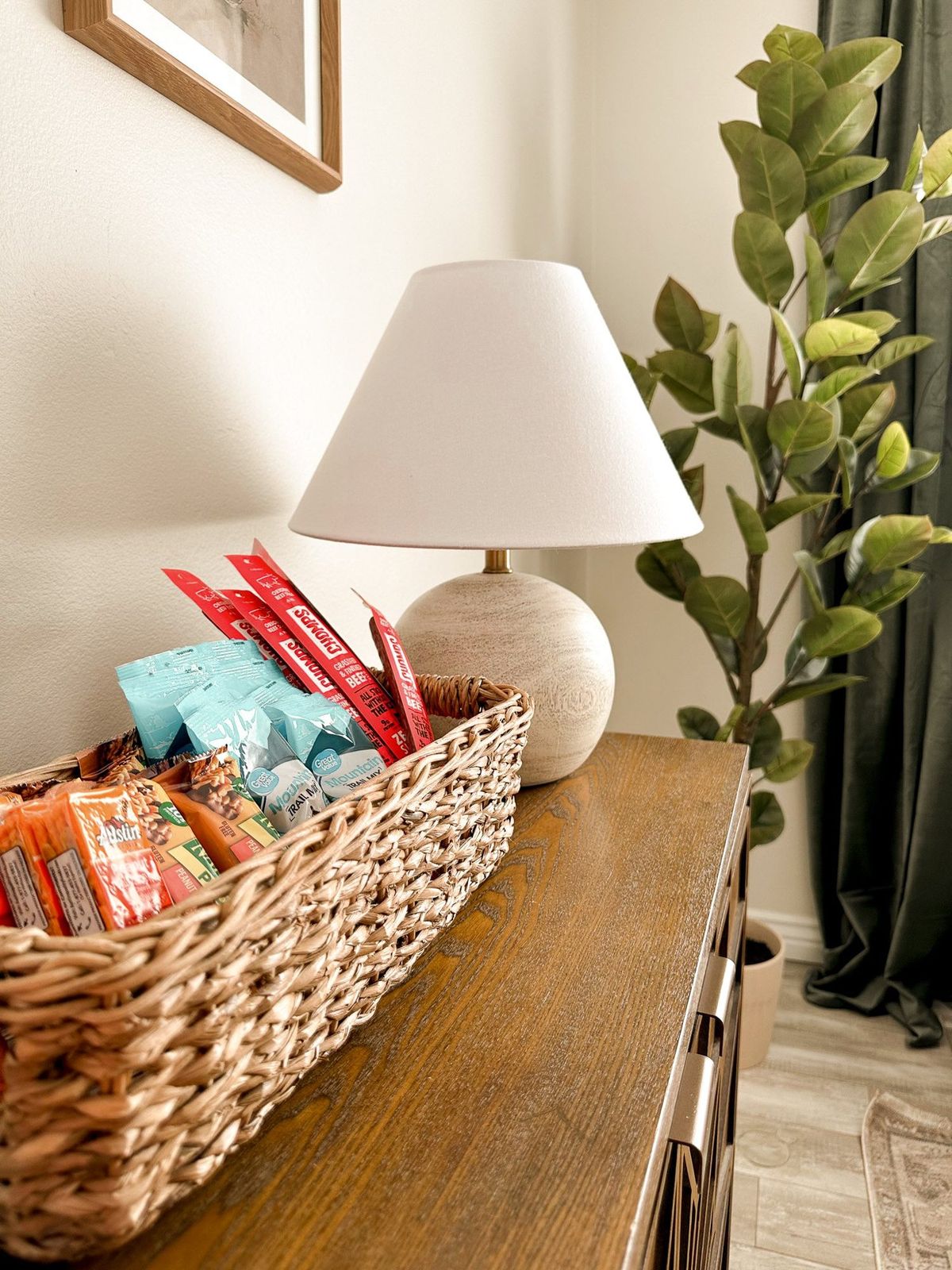 A woven basket filled with colorful snack packets sits on a wooden dresser beside a ceramic lamp. A lush potted plant is visible in the background.