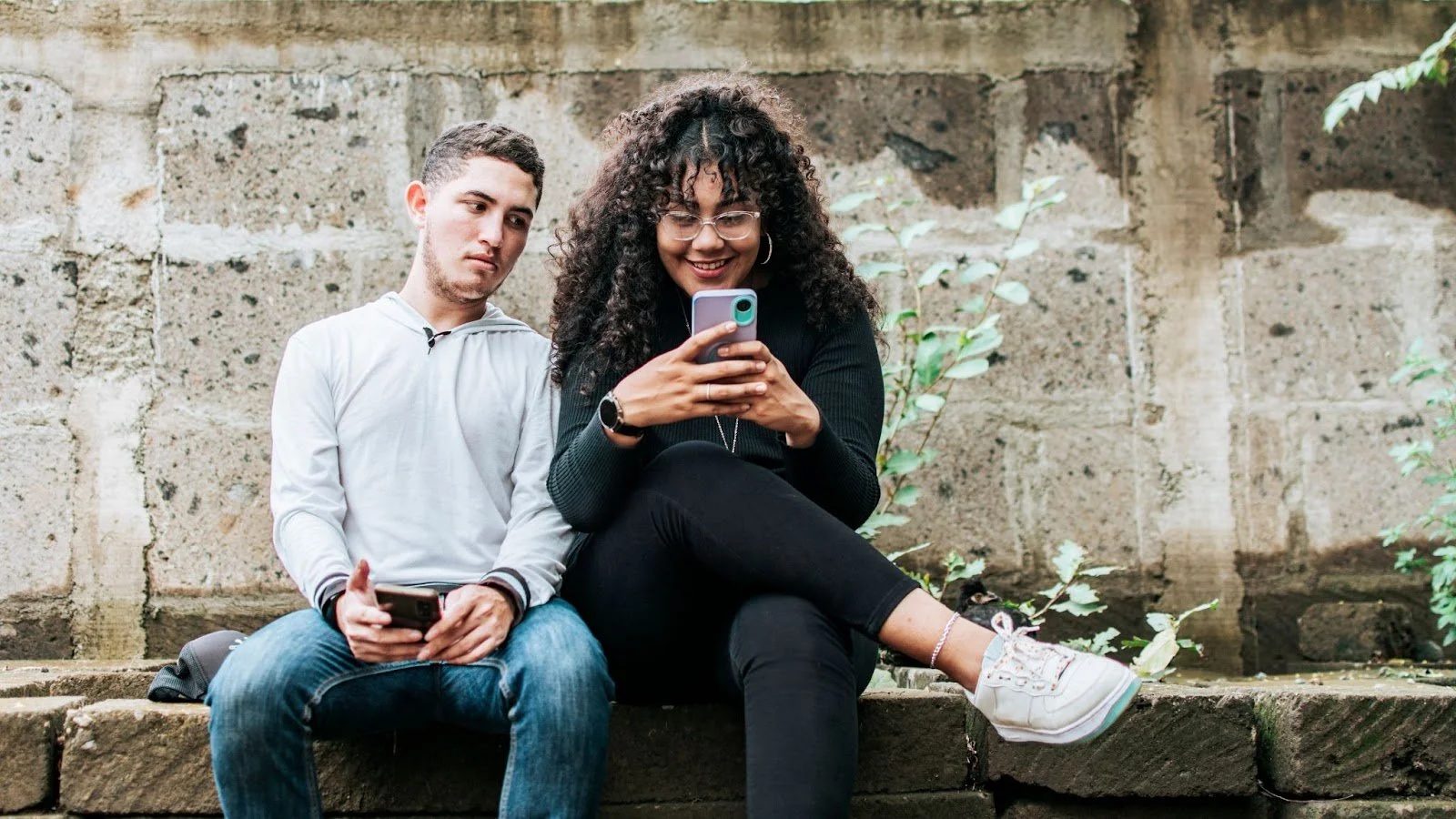 A young man and woman sitting outdoors on a stone ledge, both looking at their smartphones with a neutral to slightly smiling expression.