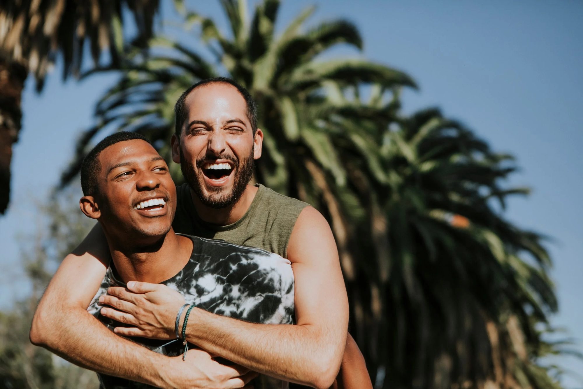 Two gay men are joyfully embracing, one behind the other, both smiling broadly. Palm trees and a clear blue sky form the backdrop, conveying a sunny, carefree vibe.