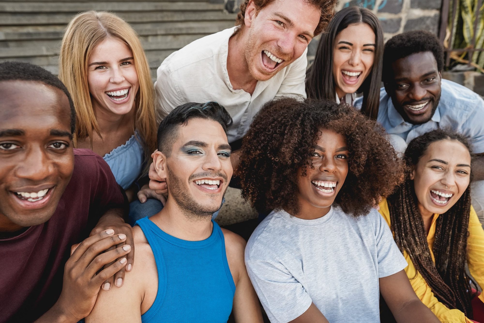 Three LGBTQ people sit closely on a rooftop, smiling brightly. They are draped in a rainbow flag, with colorful clothing, exuding a joyful, celebratory mood.