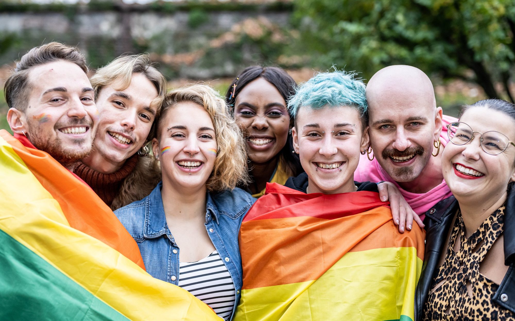 A diverse group of LGBTQ people smiling outdoors, wrapped in rainbow flags, conveying joy and unity. Trees with autumn leaves are visible in the background. Supporting LGBTQ Therapy at Tangible Therapy