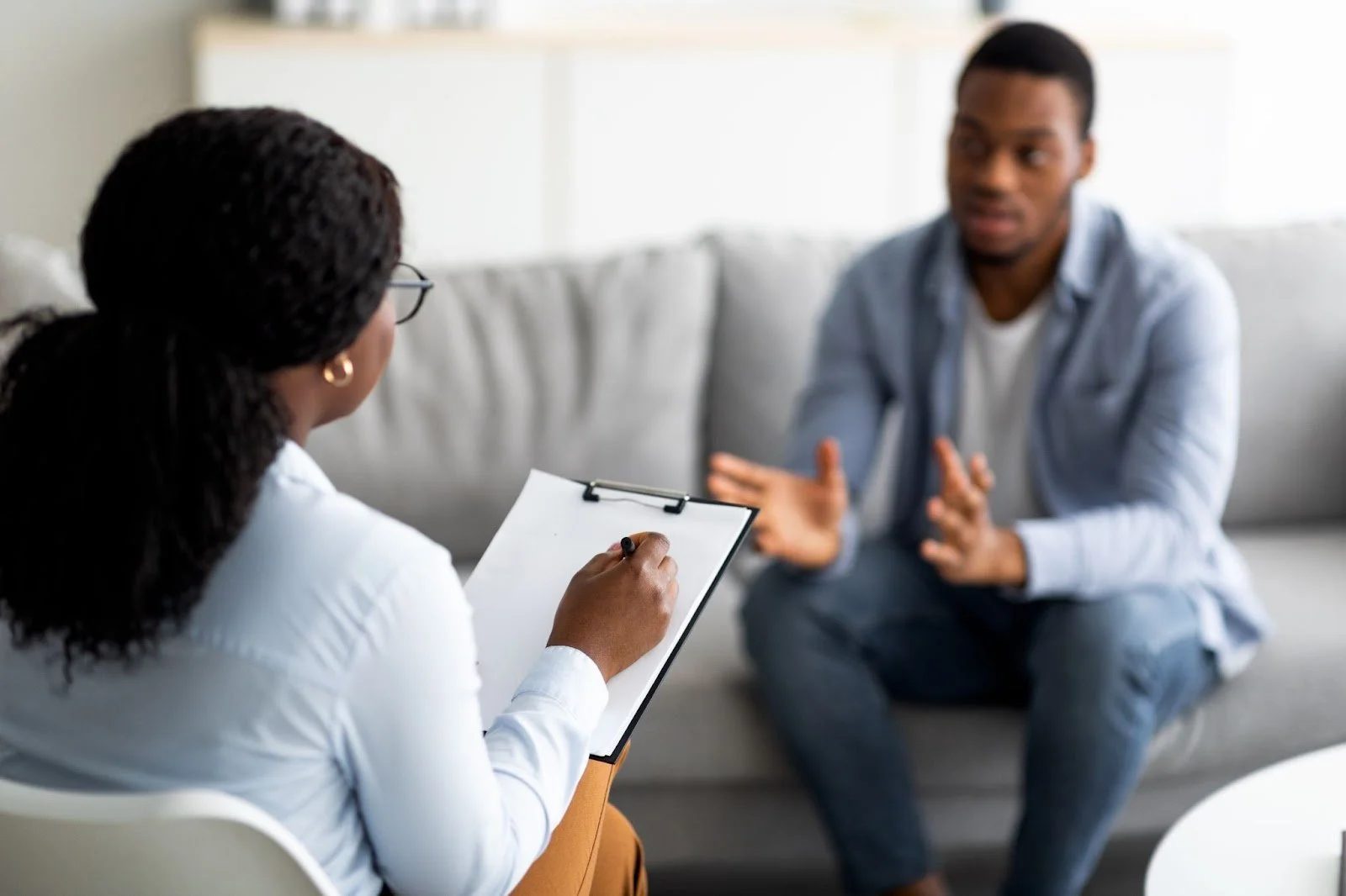 An over-the-shoulder view of a therapist taking notes on a clipboard while a man speaks during a counseling session.