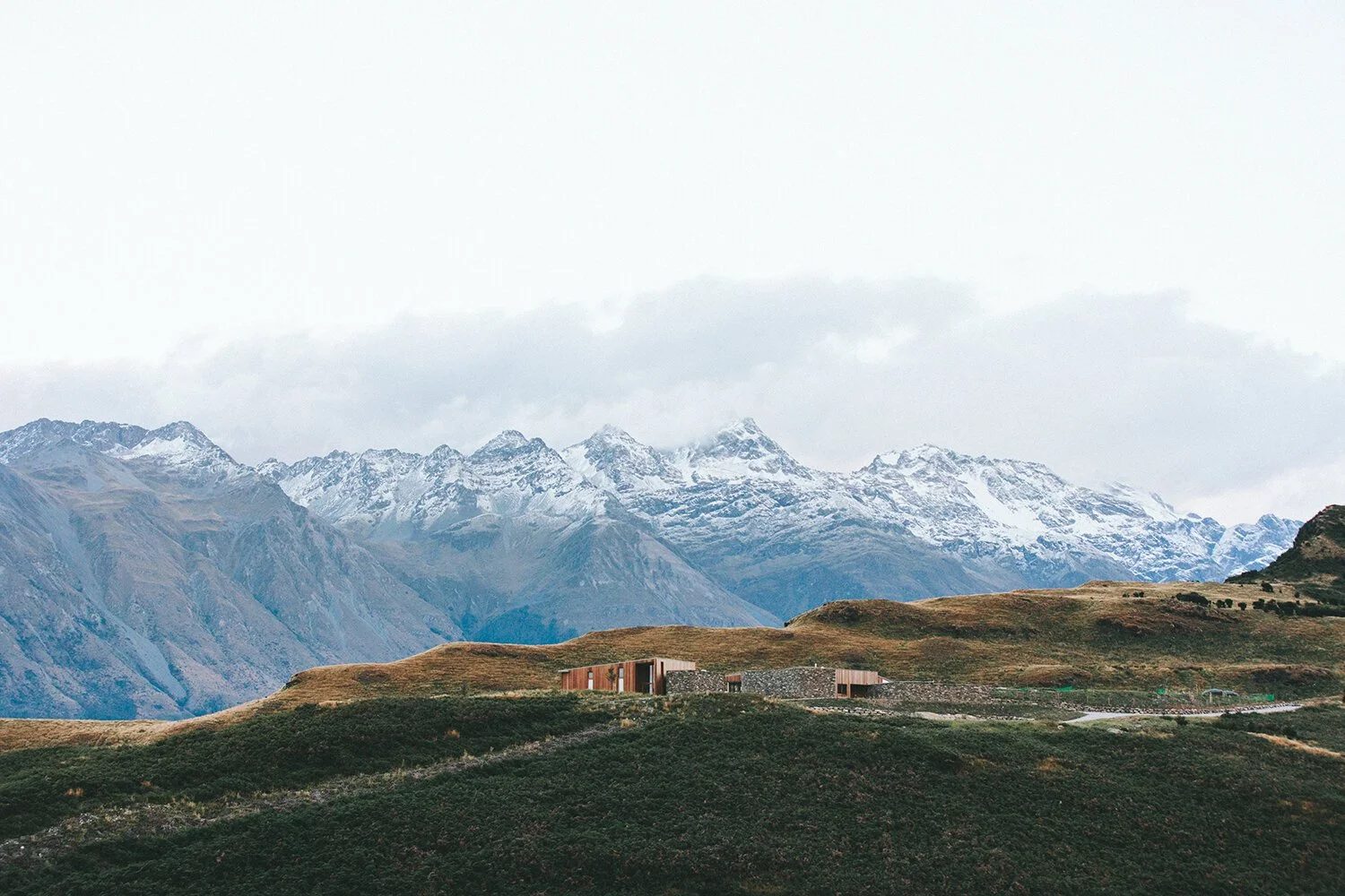 A modern house with wooden and stone exterior walls situated on a grassy hillside, with snow-capped mountains and cloudy sky in the background.