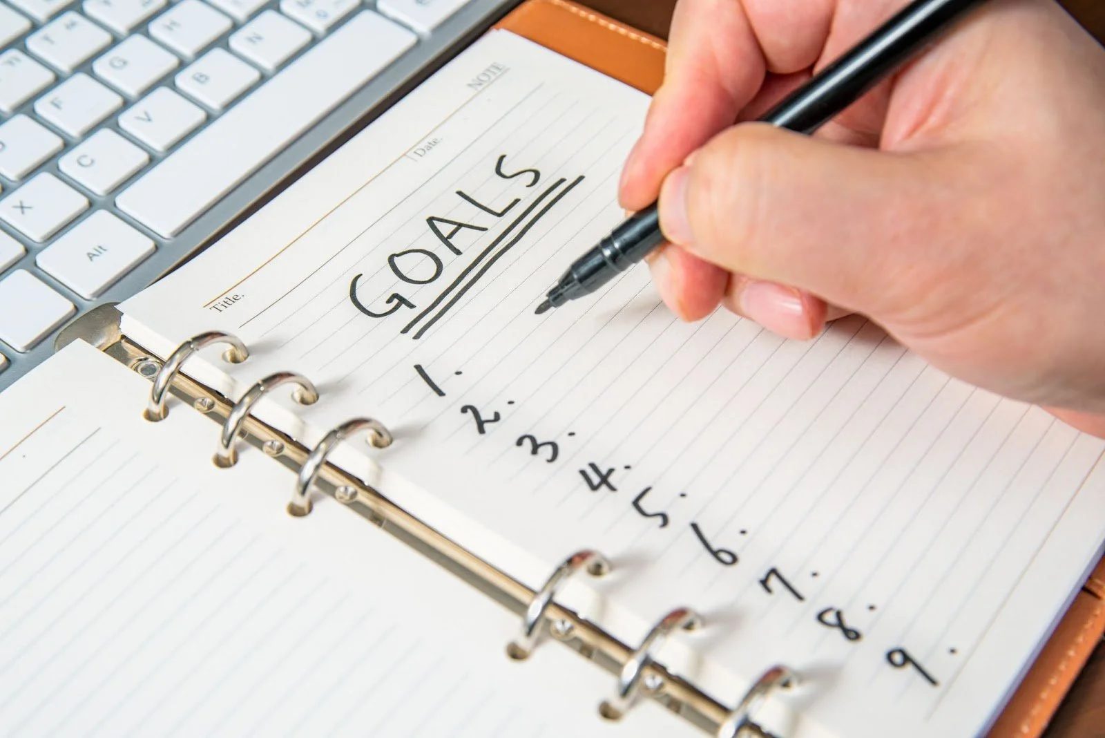 Close-up of hand writing 'GOALS' in a notebook planner with black pen next to computer keyboard for goal setting and planning