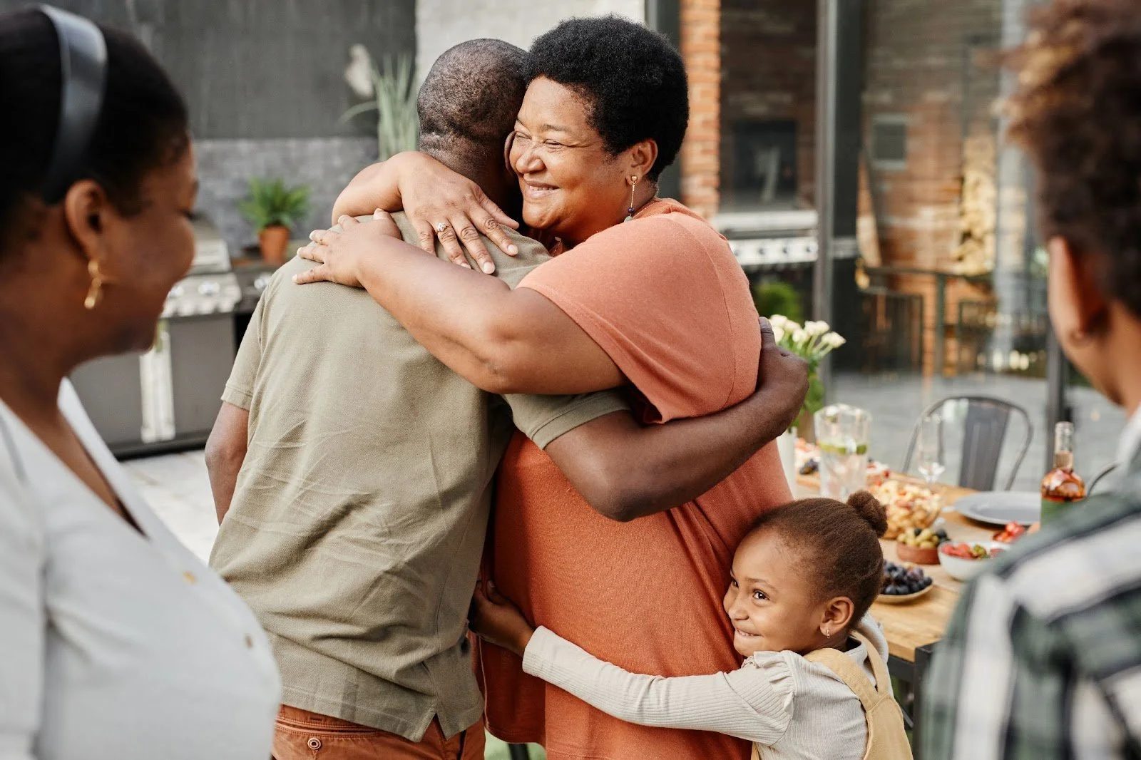 Family hugging joyfully at outdoor gathering with food table in background.