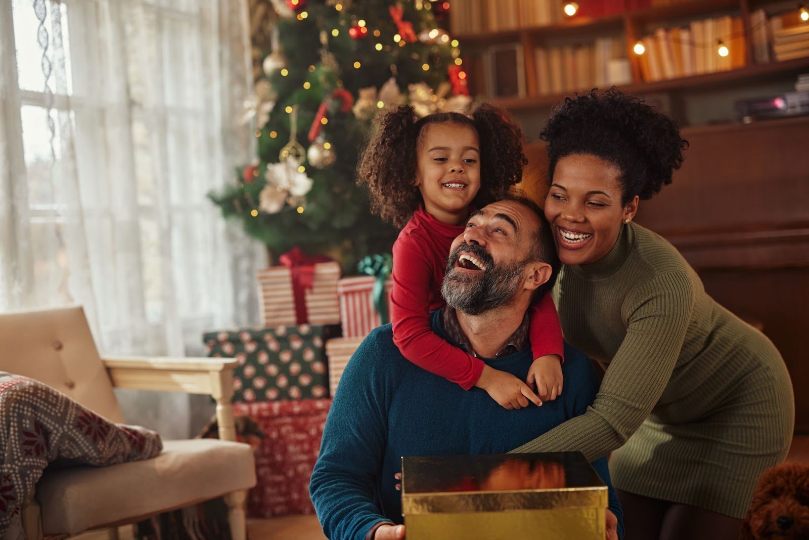 Happy interracial family laughing together by Christmas tree, father holding gift, daughter on his shoulders, mother hugging them. Cozy holiday joy.