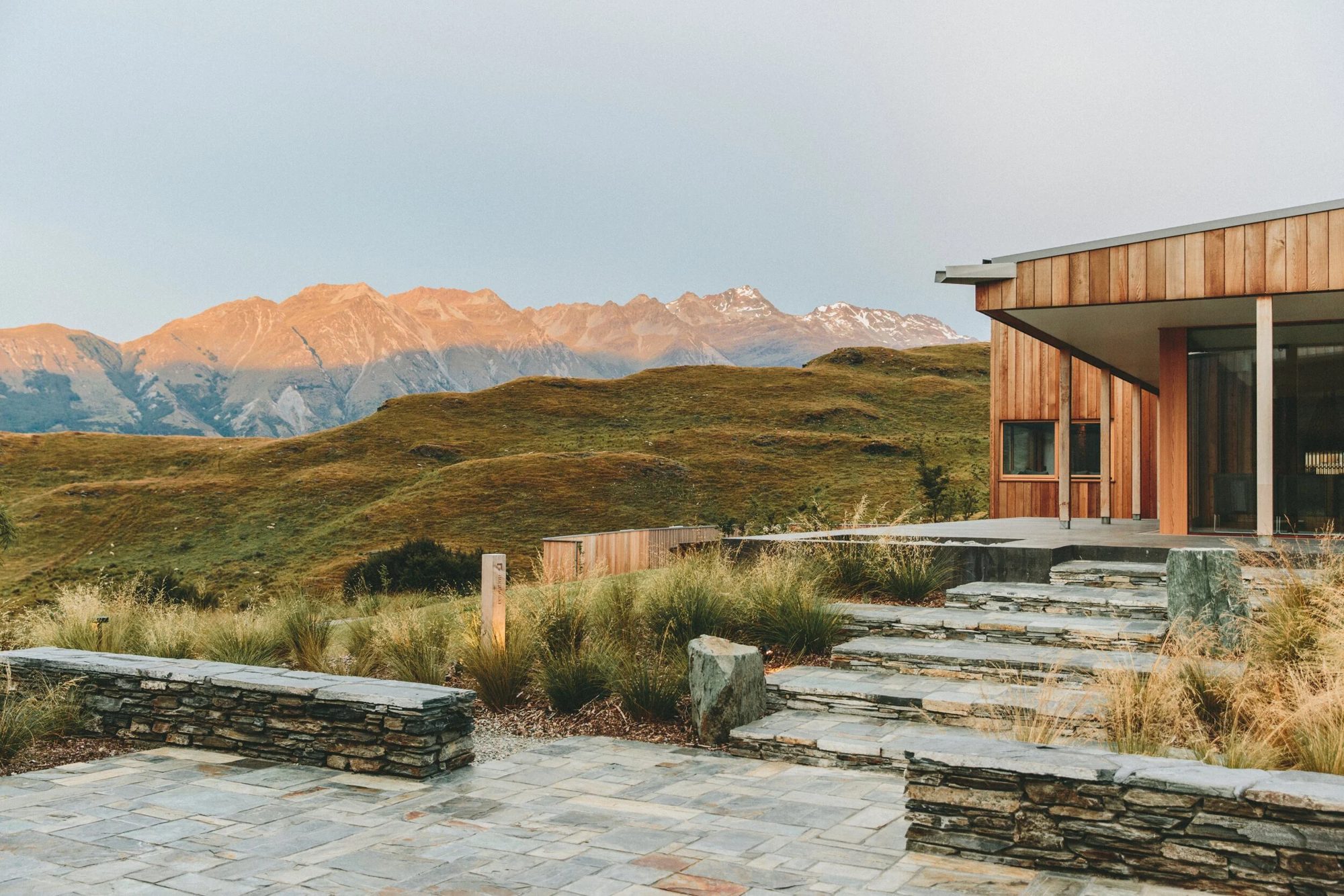 Modern wooden house on a hillside with stone steps, surrounded by grass and shrubs, mountain range in the background at sunset.