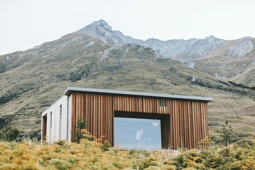 Modern house with wood and white exterior, large front window, set in a mountainous landscape with sparse vegetation.