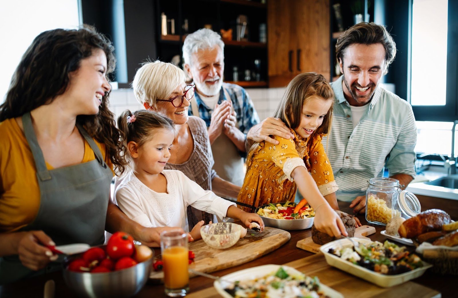 Multi-generational family joyfully cooking together in a bright kitchen, with grandparents, parents, and young children preparing food and smiling.