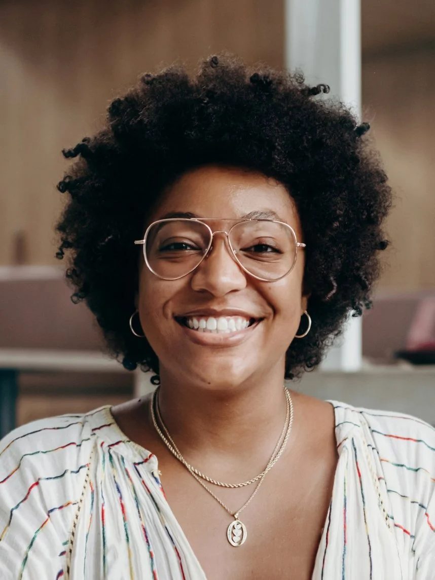 Smiling woman with curly hair, glasses, hoop earrings, layered necklaces, and a striped shirt in an indoor setting.