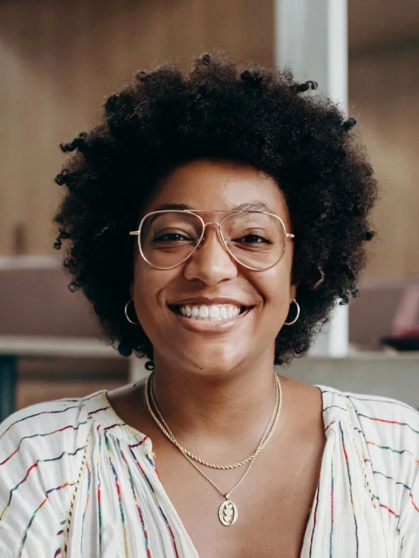 Smiling woman with curly hair, glasses, hoop earrings, layered necklaces, and striped blouse.