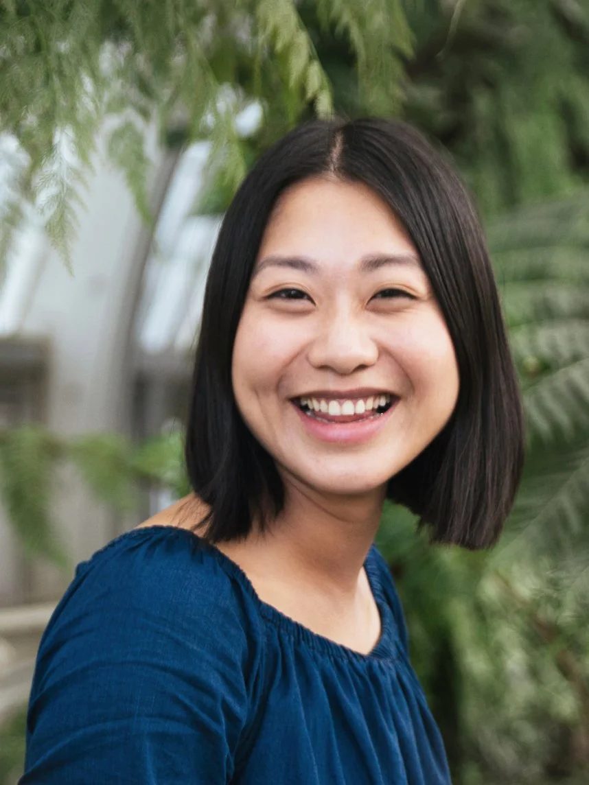 A smiling woman with shoulder-length black hair, wearing a blue top, standing outdoors with green plants in the background.