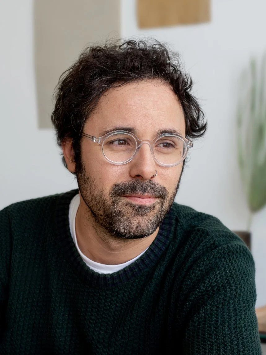 A man with curly dark hair, glasses, and facial hair, wearing a black sweater, sitting indoors with a neutral background.