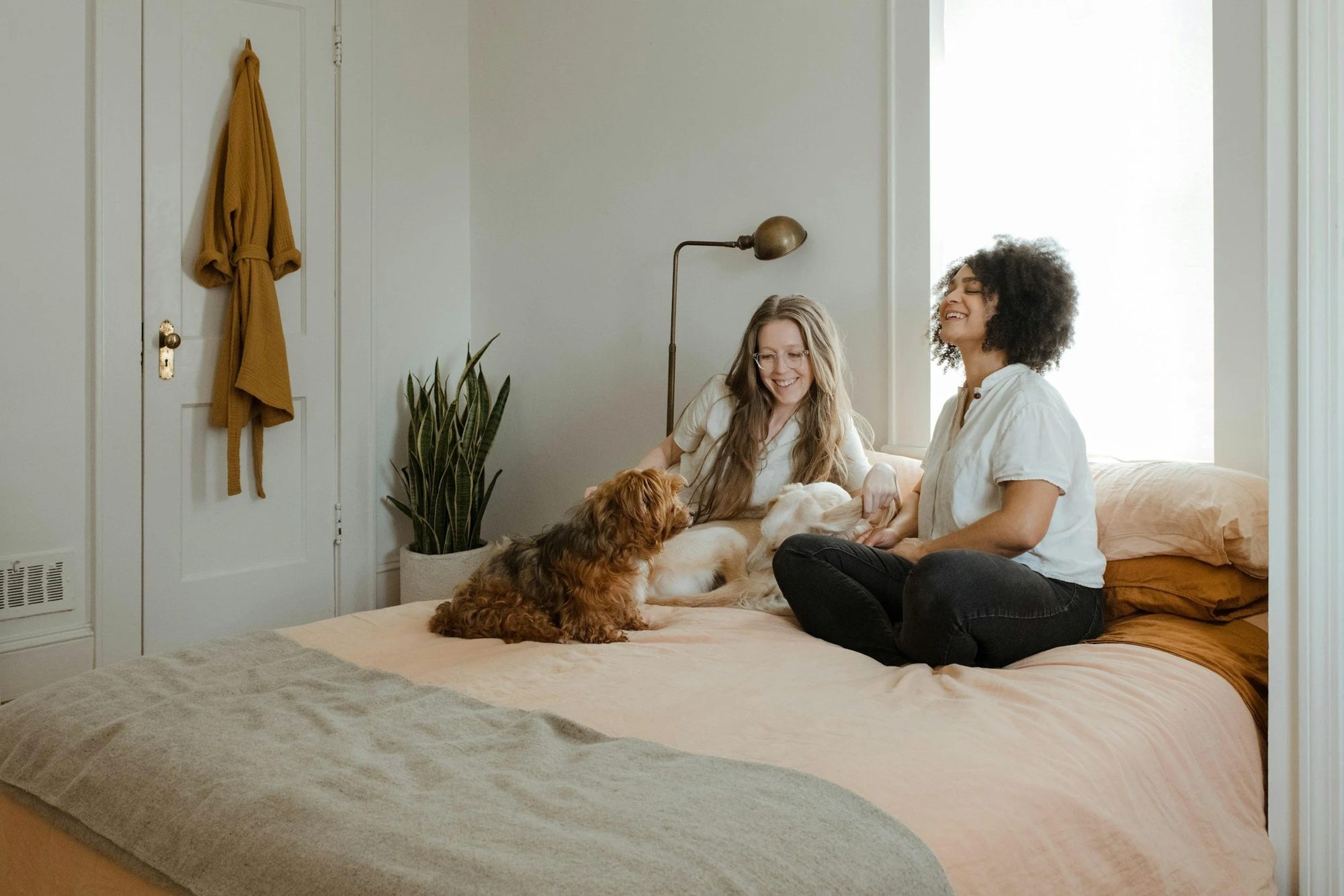 Two women with relational OCD sit on a bed, smiling and petting two small dogs. The room is cozy with soft lighting, light-colored bedding, and a houseplant.